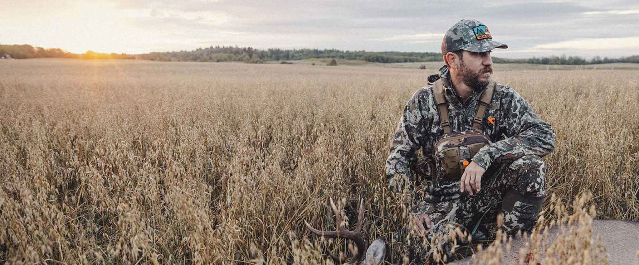 Hunter in multi-camo apparel with tan chest rig kneeling near harvested deer.