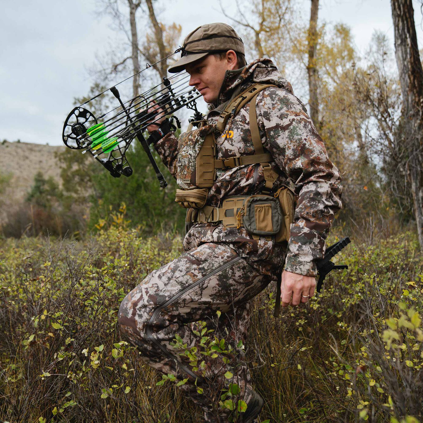 Camo hunter in multi-pattern suit with side zip pants, holding compound bow & green arrows.