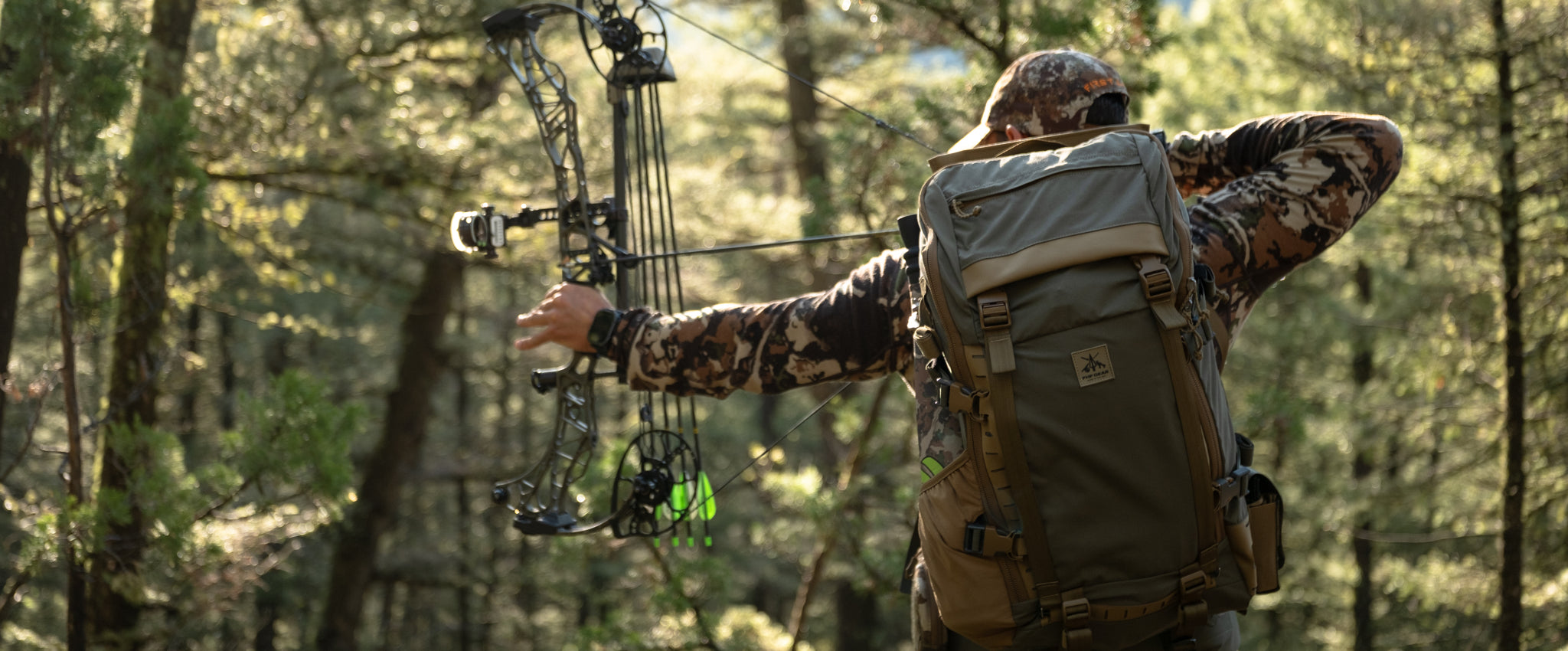A person in camouflage clothing and a large olive green backpack draws a compound bow in a wooded forest setting.