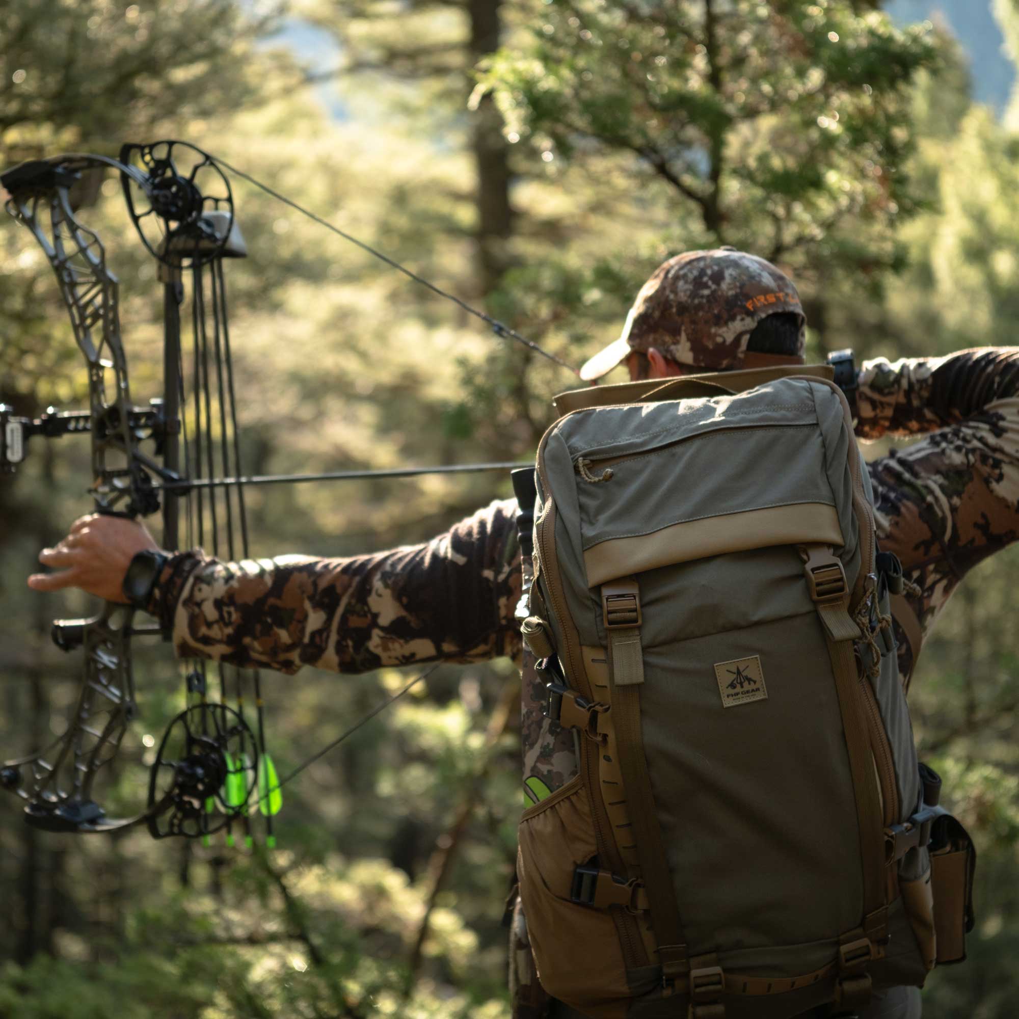 A person in camouflage clothing and a cap stands outdoors in a forest, holding a compound bow with a large olive-tan backpack on their back.