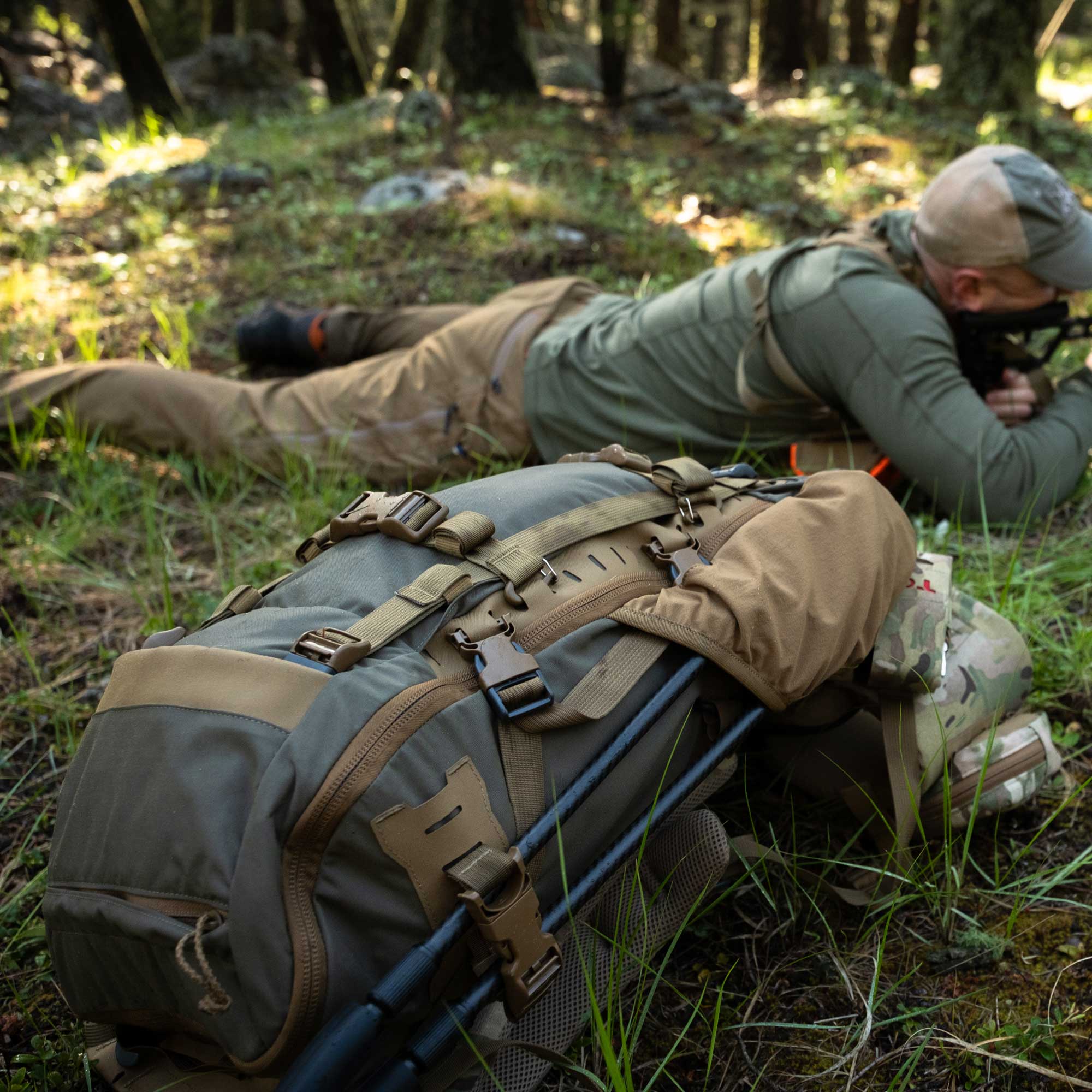 A large olive green and tan backpack with trekking poles resting on its front, set on a grassy forest floor next to a person in camouflage clothing lying prone in a wooded outdoor environment.