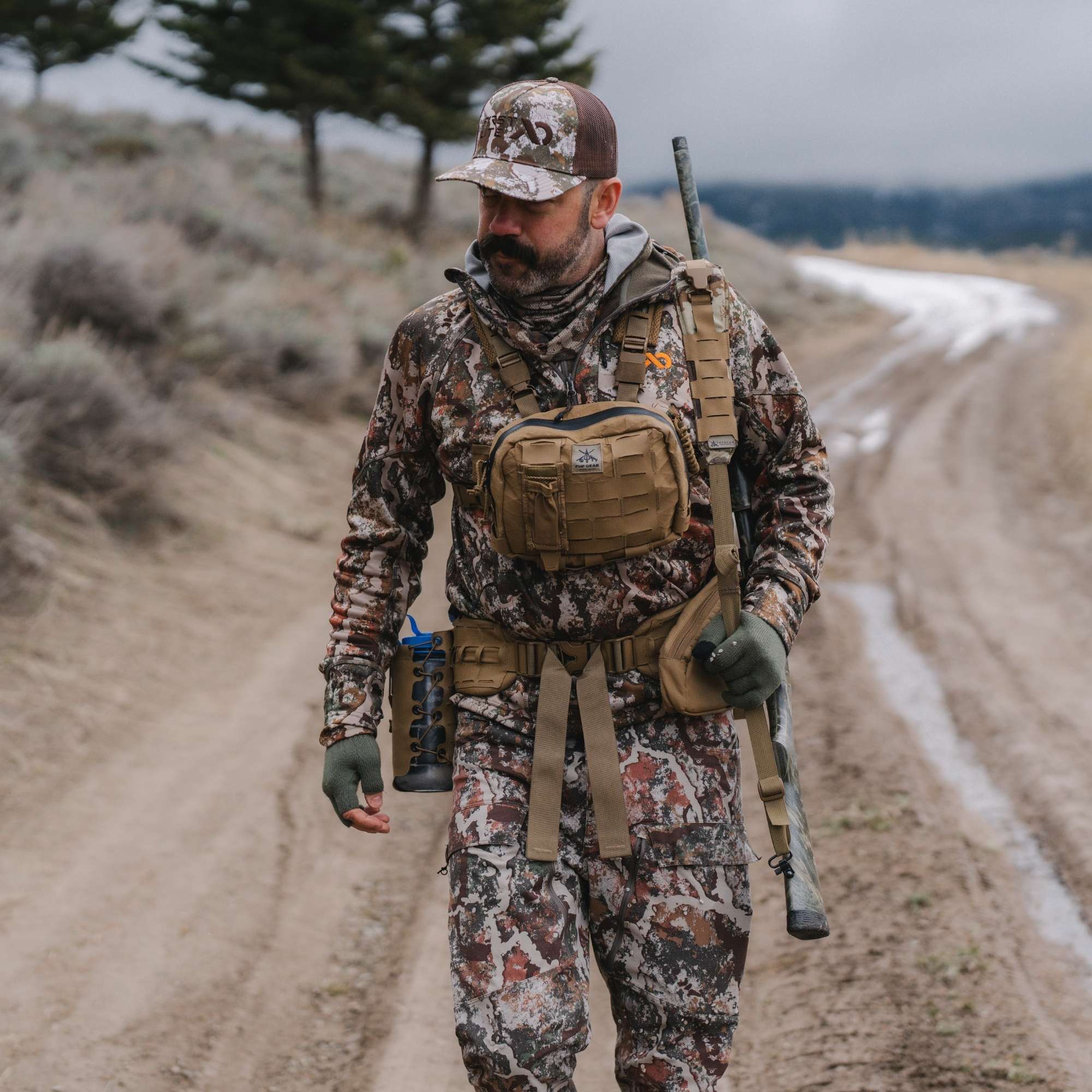 Man in the field dressed in camo gear, a water bottle, a rifle and a chest rig