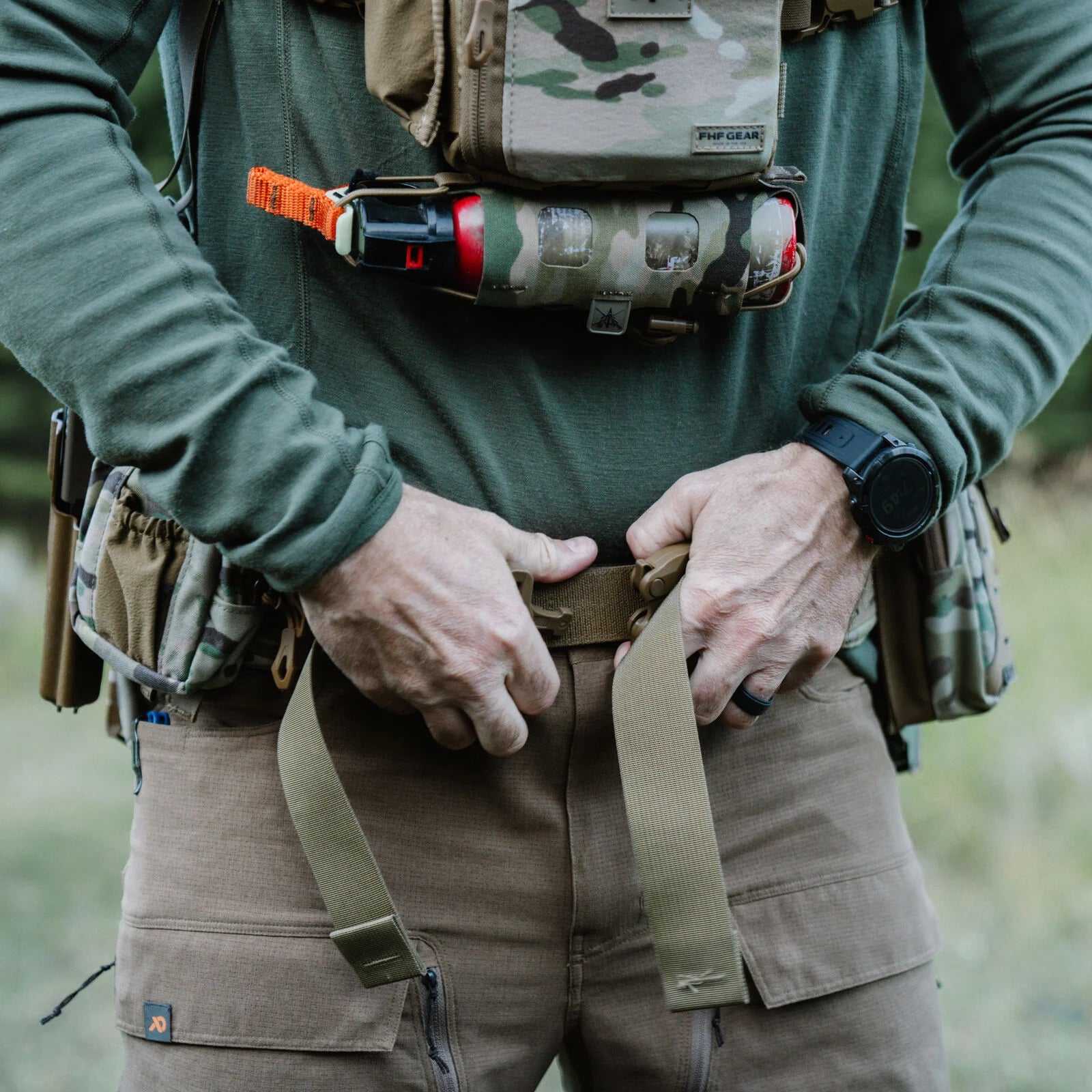 Brown tactical pants, tan belt being adjusted, olive shirt, and camo chest rig with canister.