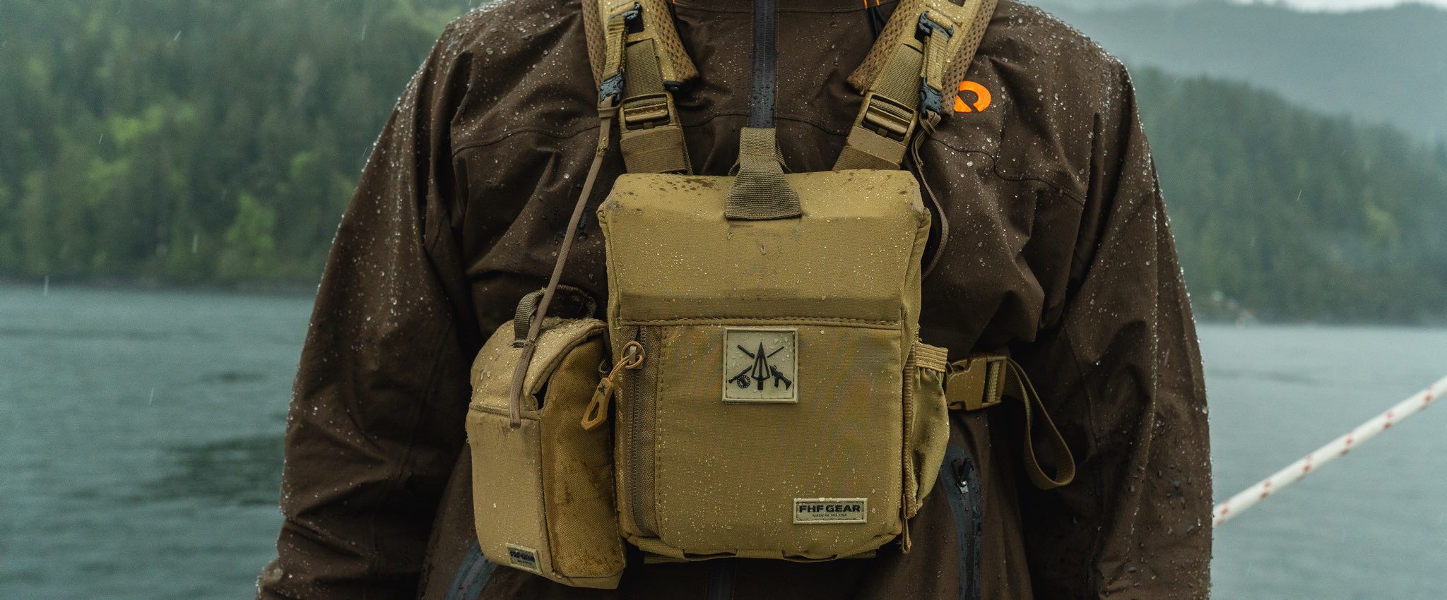 A tan bino harness worn by a person in a wet brown waterproof jacket, with a lake and forested hills in the background.