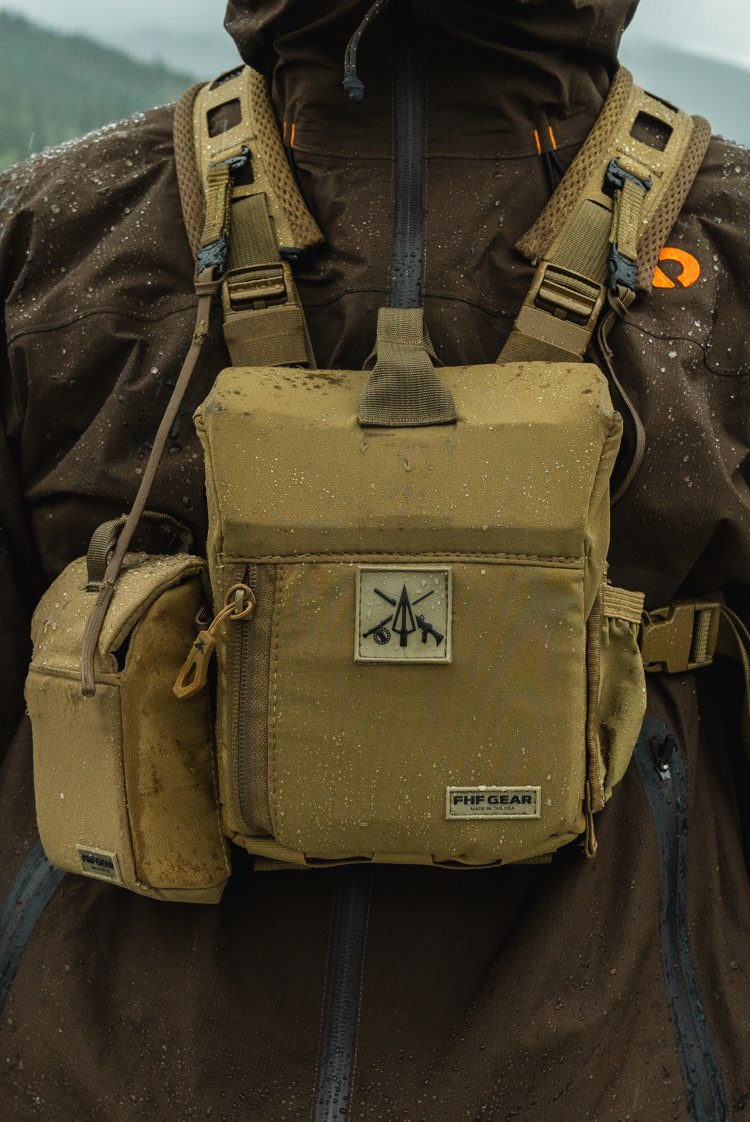 A tan bino harness worn over a brown waterproof jacket, featuring a front zip pocket with a logo patch and a side pouch, photographed outdoors in the rain.