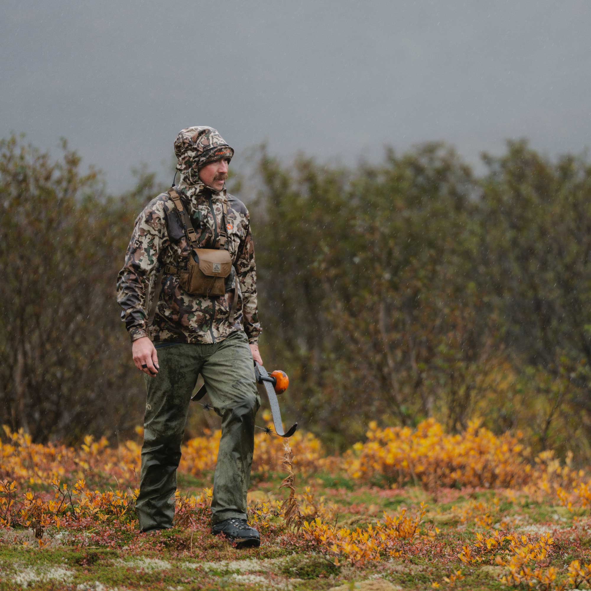 A man wearing a camouflage hooded jacket, green pants, and a small brown chest pouch walks through a grassy, shrub-filled outdoor landscape holding a bow.