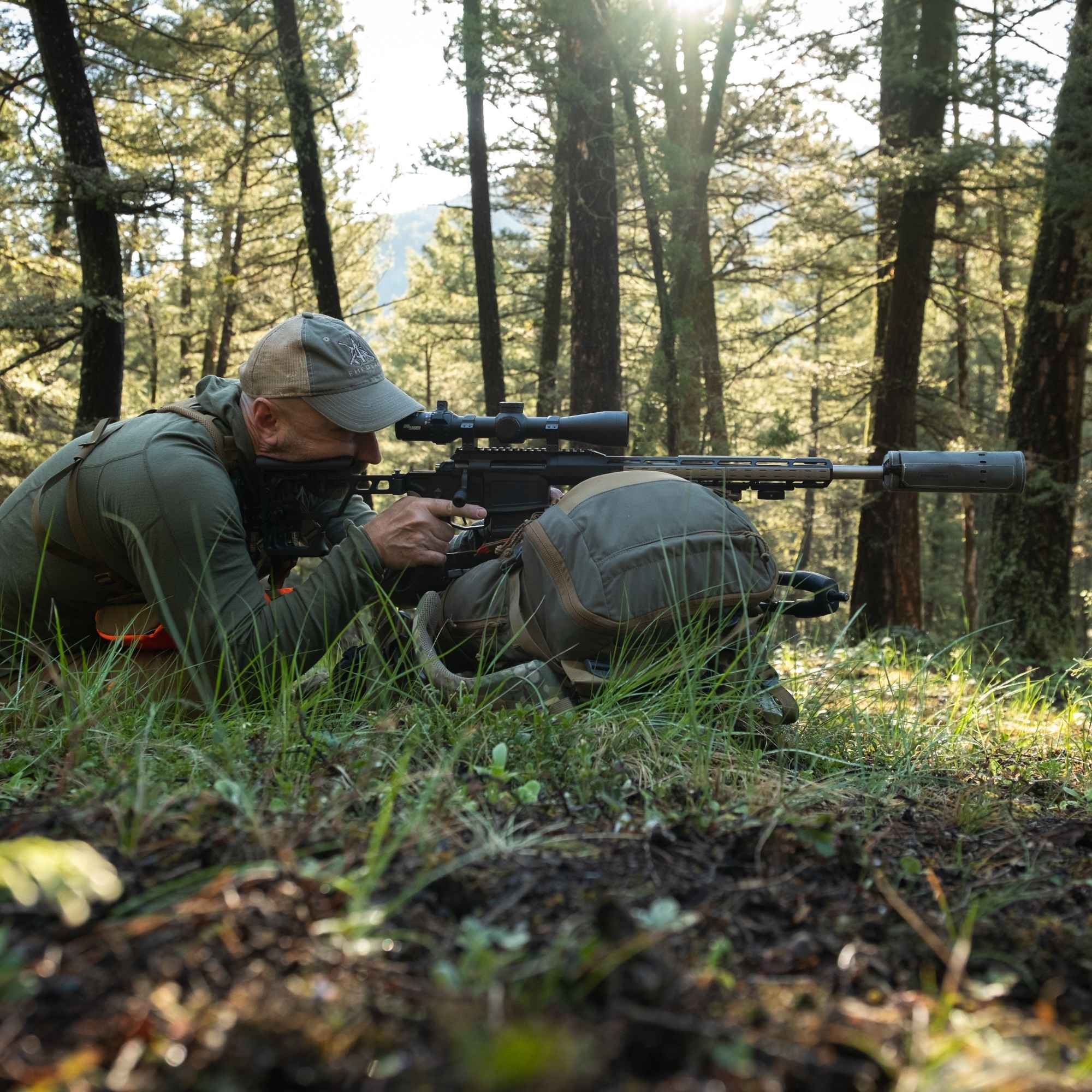 Hunter aiming rifle with black scope on a green bag, green suppressor cover in a forest
