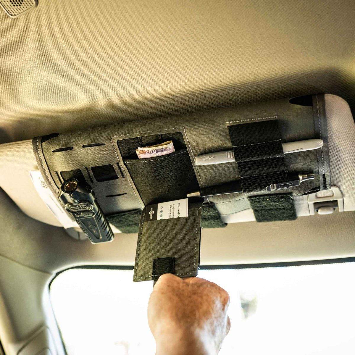 Hand pulling a patch of a gray car visor organizer that has cards in it and has other objects in the remaining storage options.