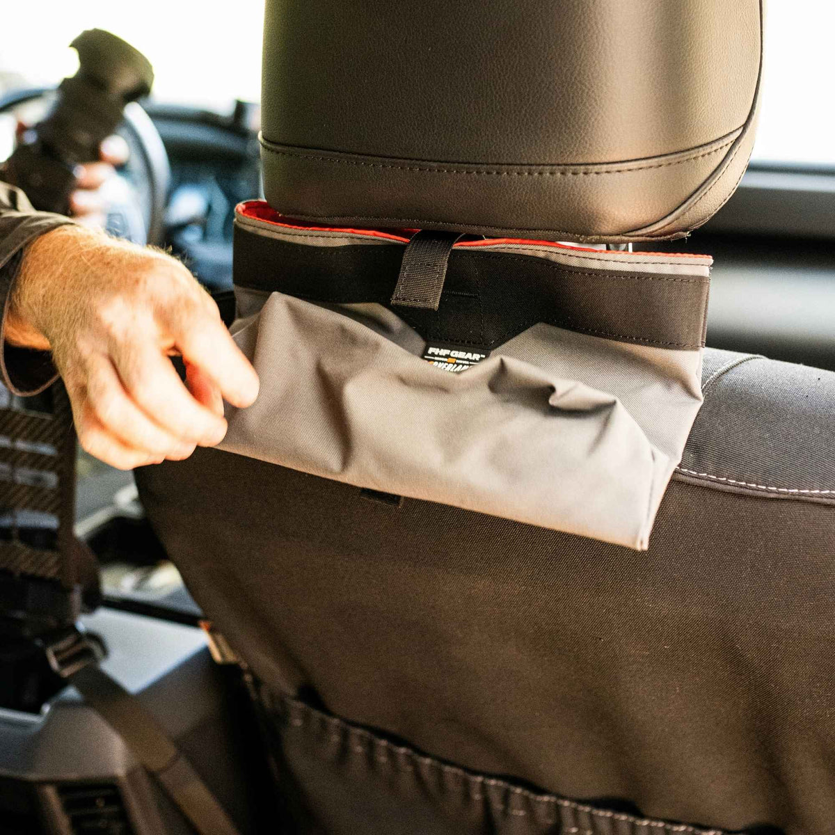 Man unrolling a grey storage bag with FHF XOVERLAND branding on the center that is mounted at the back of a headrest.
