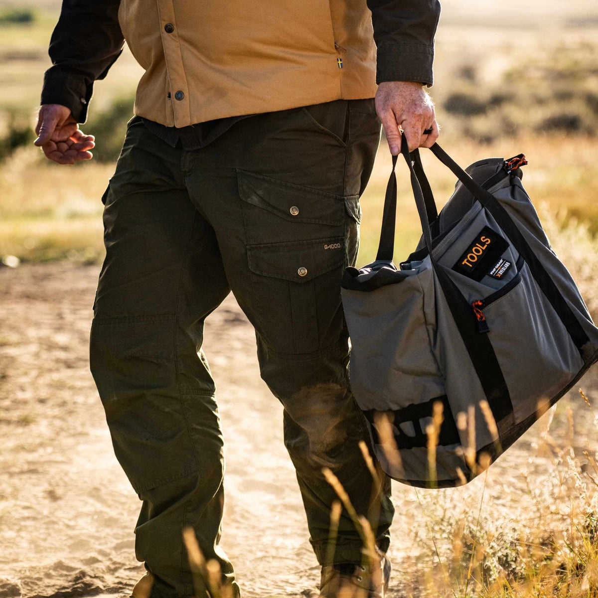 Man carrying an FHF The Stage Tote in grey with black straps, orange cords on zippers and FHF XOVERLAND branding.