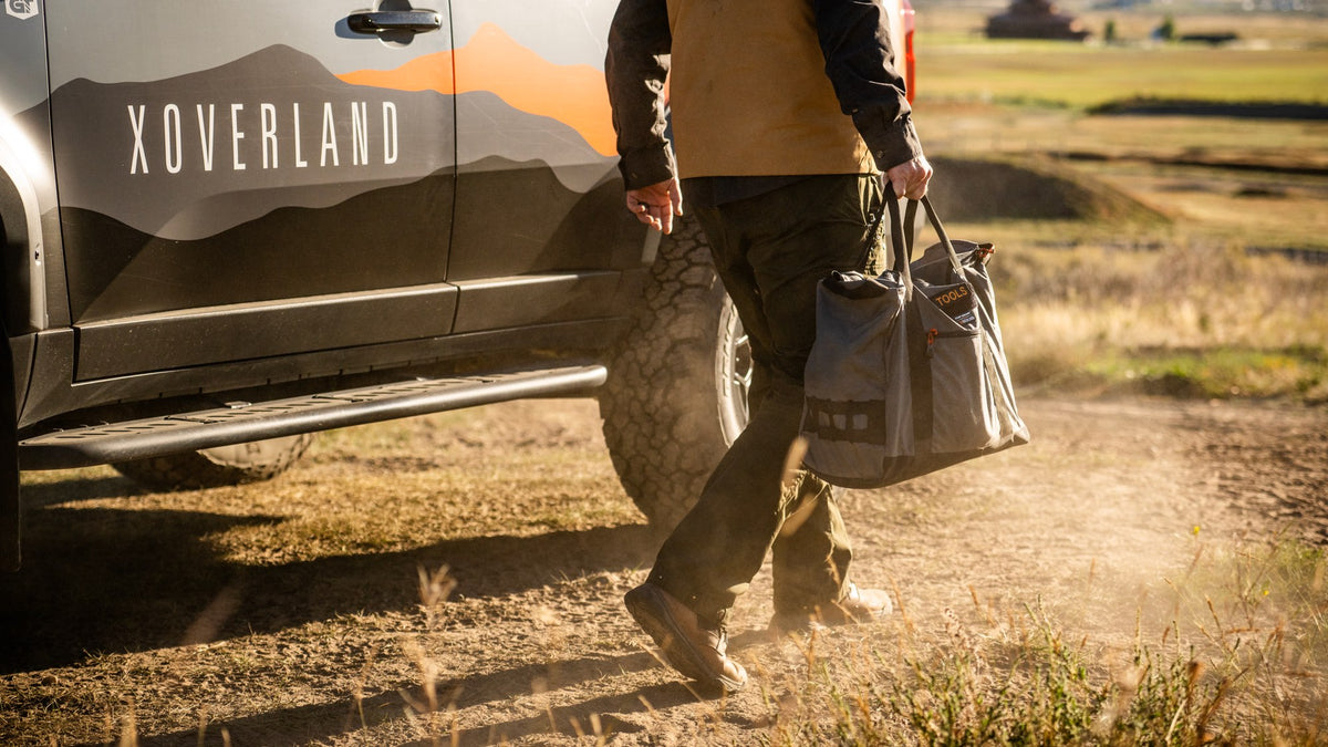A person walking on a dirt road beside a dark pickup truck with XOVERLAND branding, carrying a large gray tote bag outdoors in a rural setting.