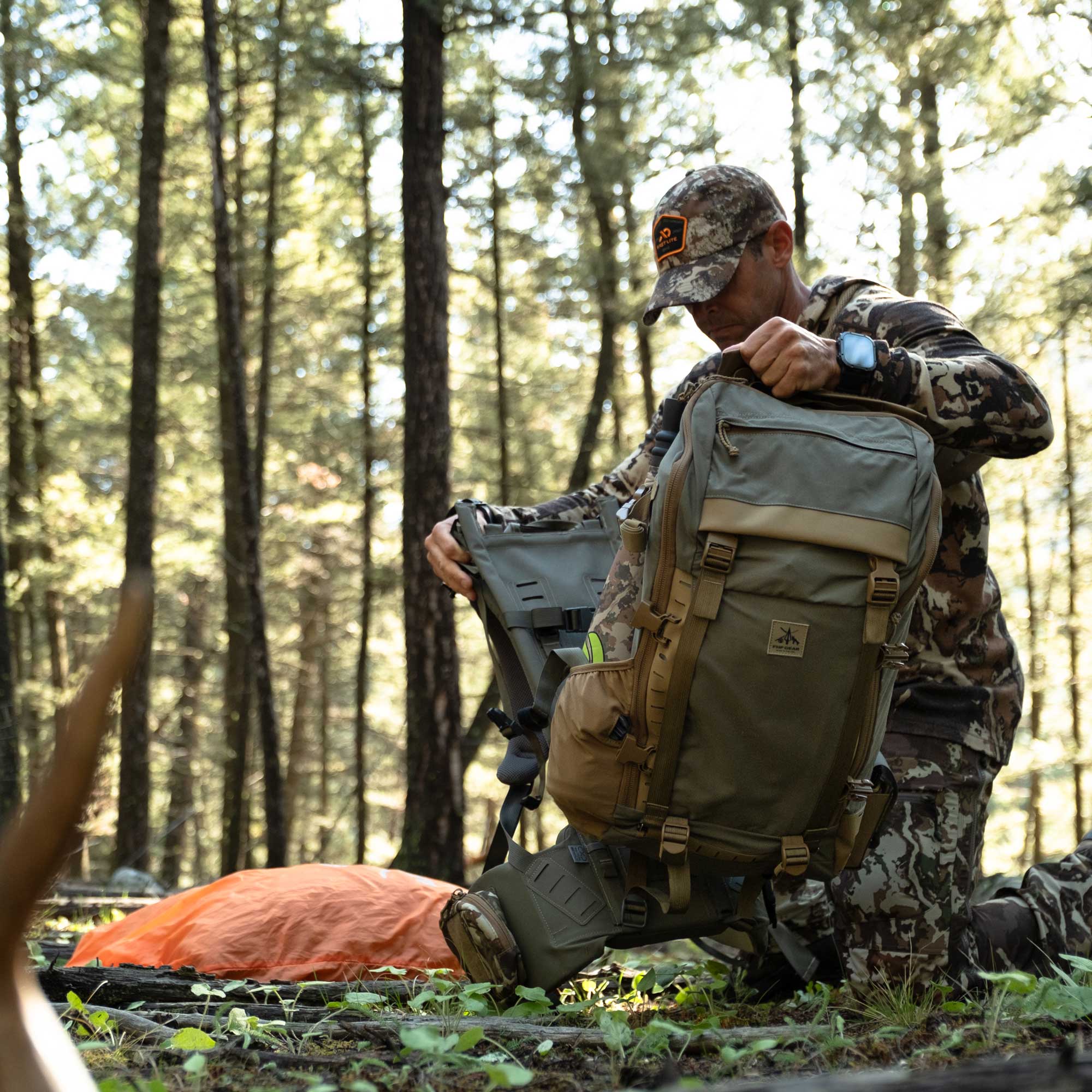A man in a camouflage hunting jacket and cap packs a large olive green and tan backpack filled with gear in a dense forest.