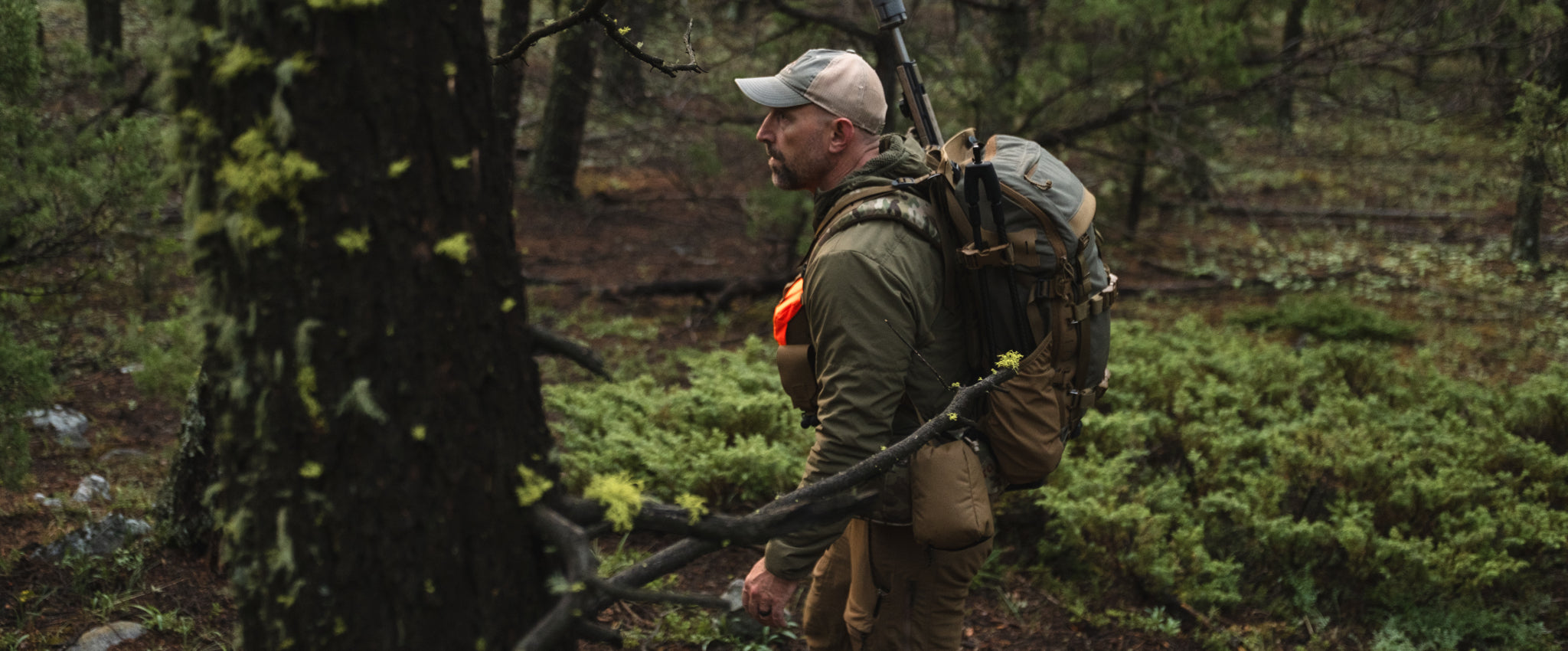 A man in outdoor hunting attire, including a camouflage cap, green jacket, and a large brown backpack, standing in a mossy forest next to a tree trunk.
