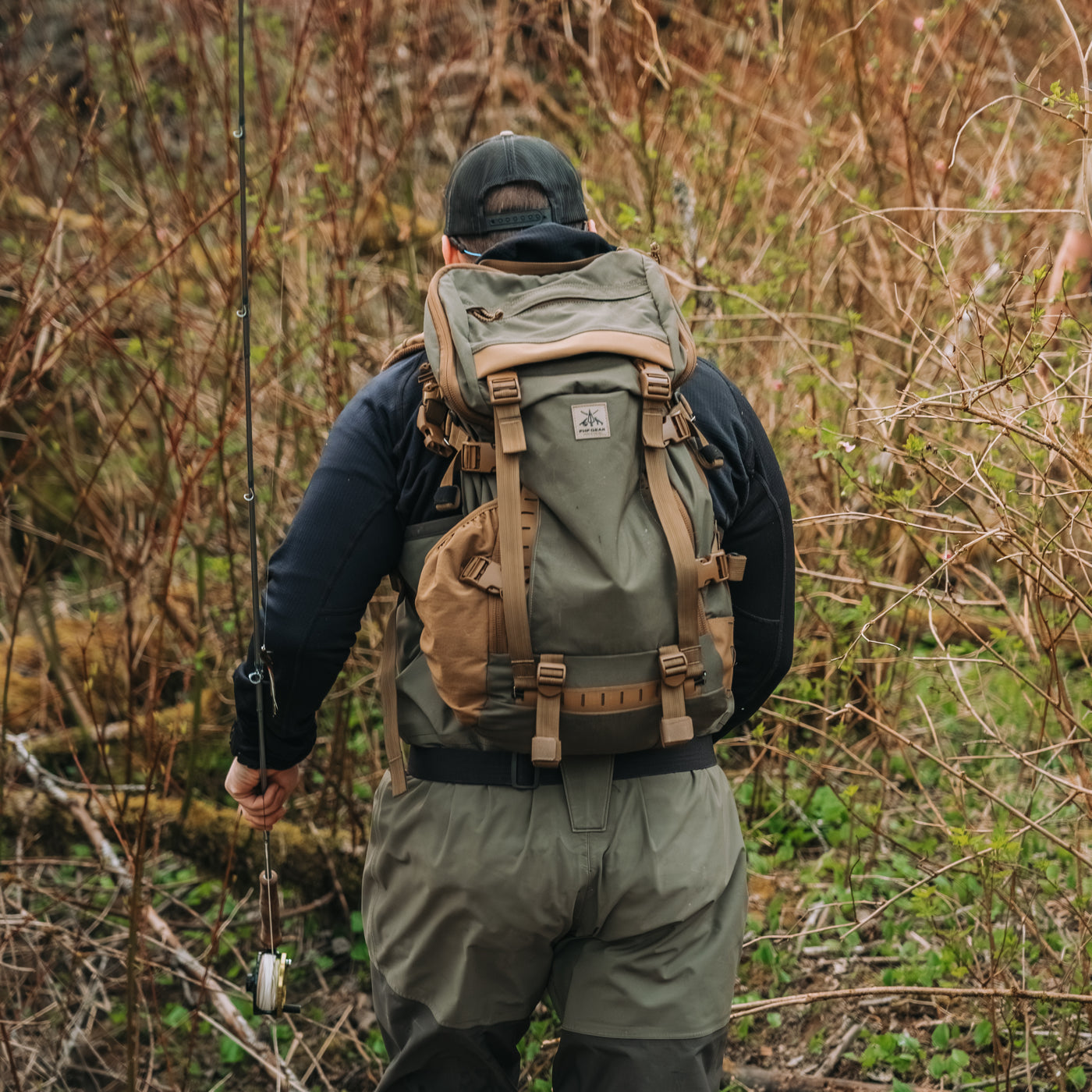 A man seen from behind wearing an olive green and tan fly fishing backpack, waders, and a black long-sleeve shirt, standing in a wooded outdoor environment holding a fishing rod.