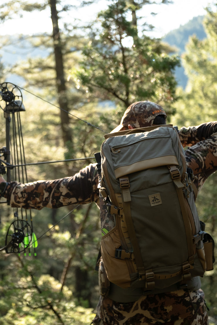 A person in camouflage clothing holds a compound bow while wearing a large olive green and tan backpack outdoors in a forest setting.