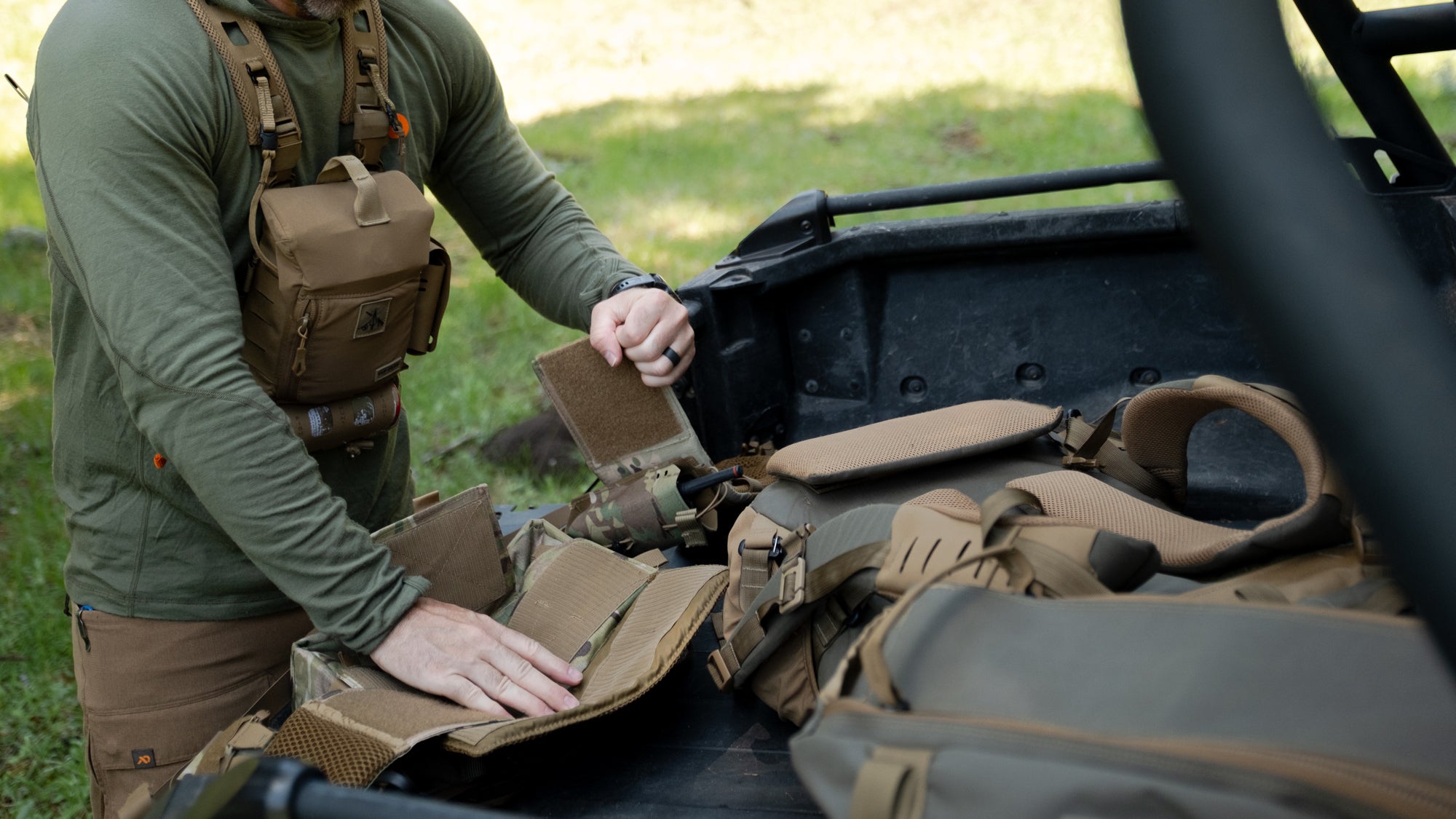 A person in an olive green shirt and tan tactical vest packs gear into the bed of a black truck outdoors.