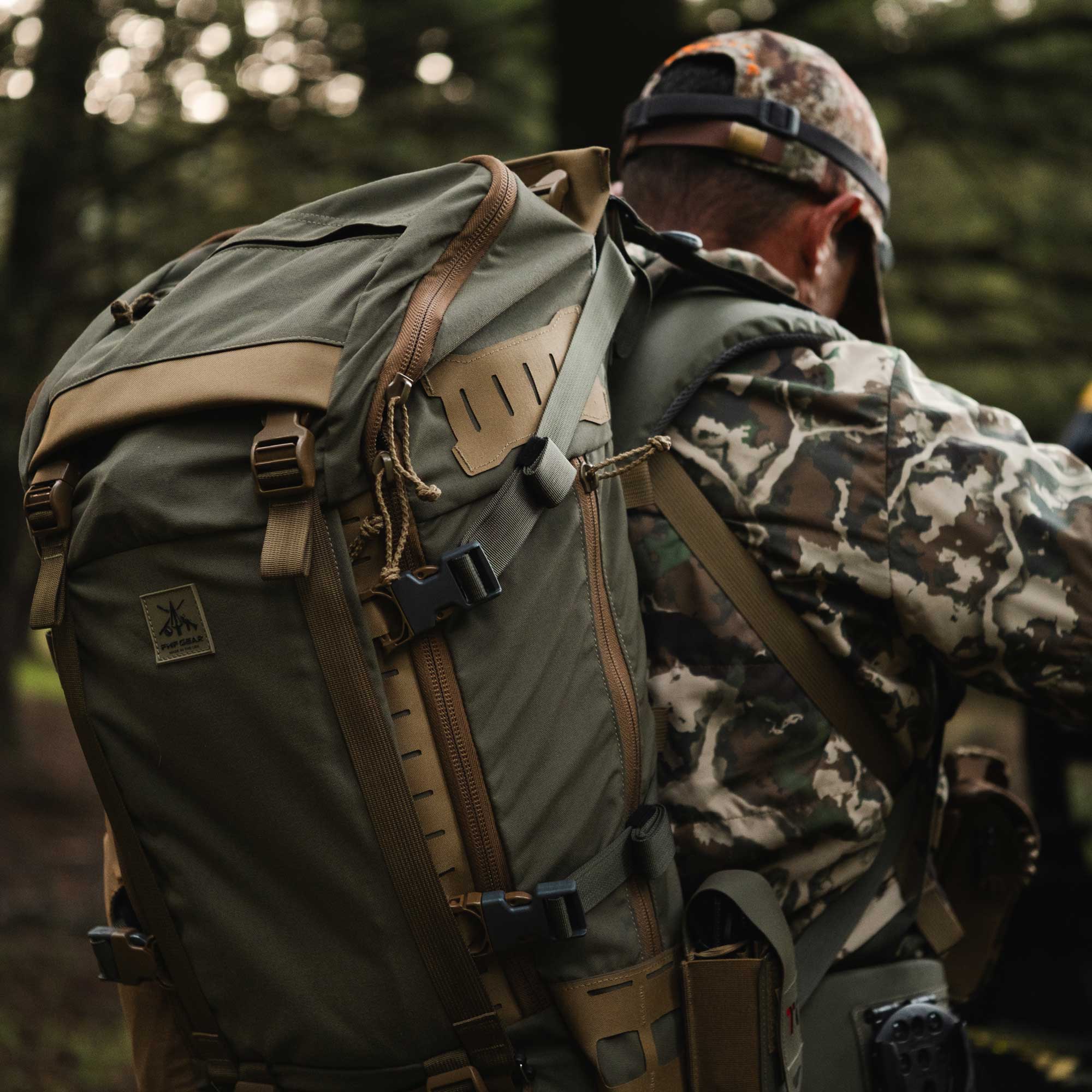 A man wearing a camouflage hunting jacket and cap, seen from behind, carrying a large olive green hunting backpack with tan straps and buckles in a forest setting.