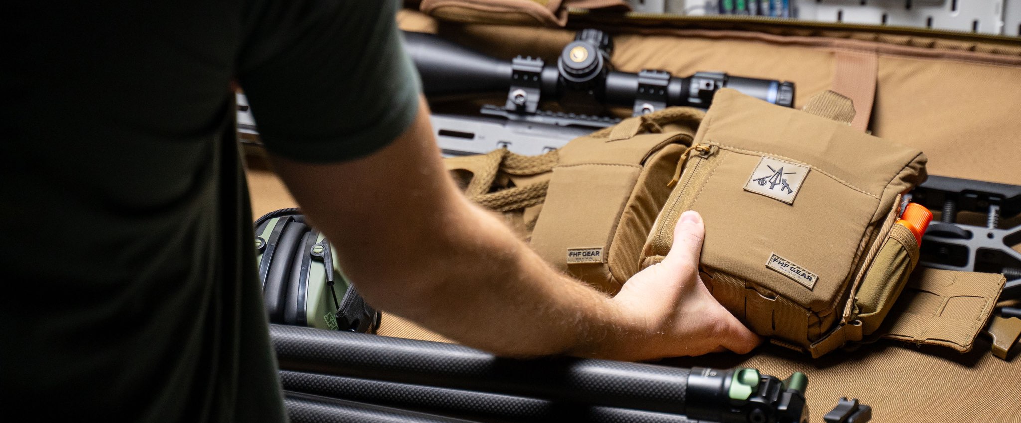 Person holding a tactical pouch with rifles and equipment in the background