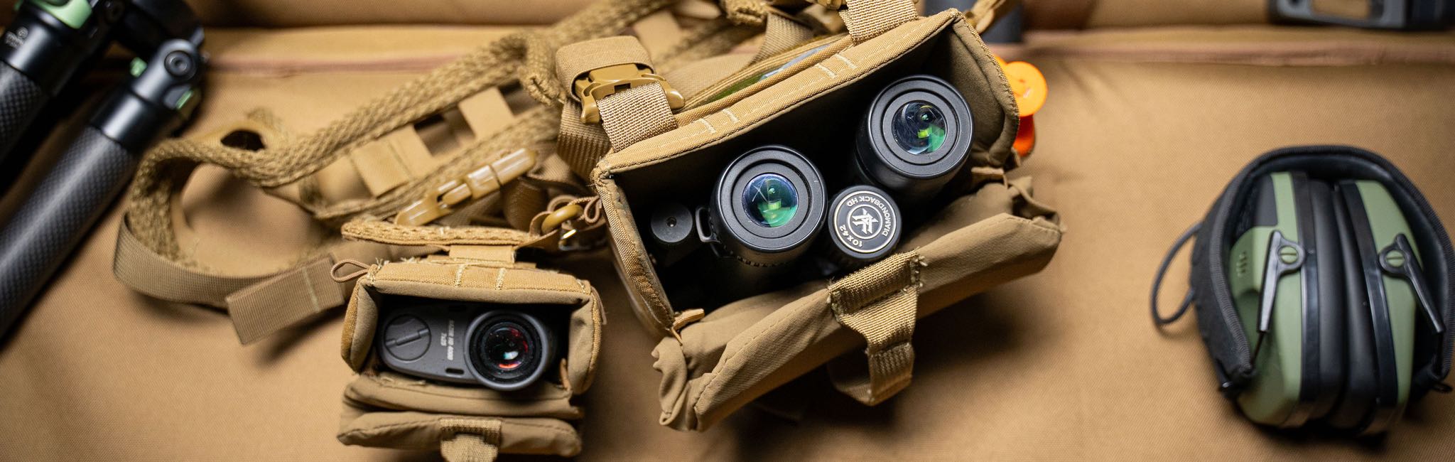 Tan tactical gear setup: binoculars, rangefinder, tripod, and green shooting ear muffs.
