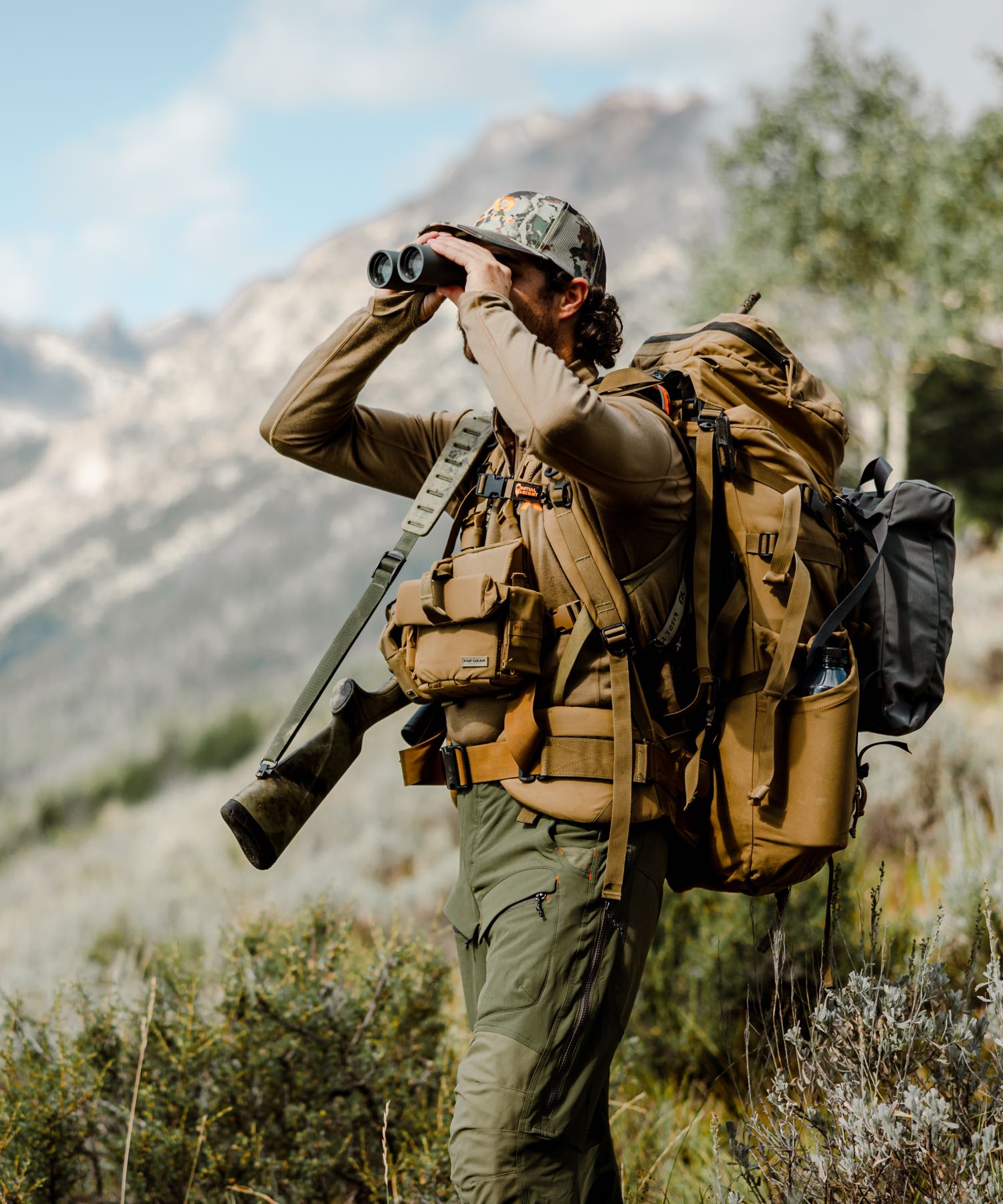 Man in tan hunting pack system & olive pants, with rifle, using binoculars.