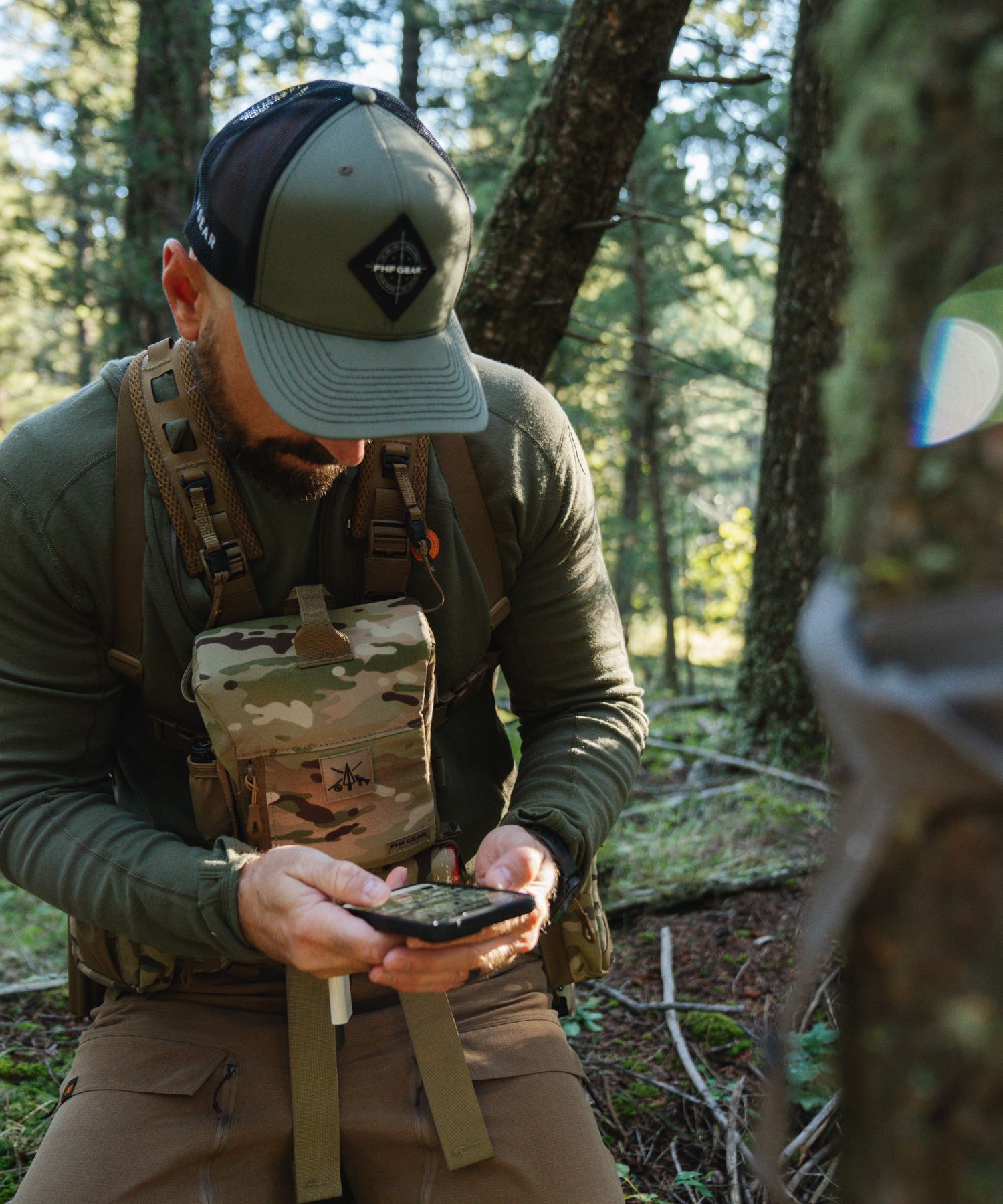 Man wears FHF Gear multicam chest pack and olive cap, looking at smartphone.
