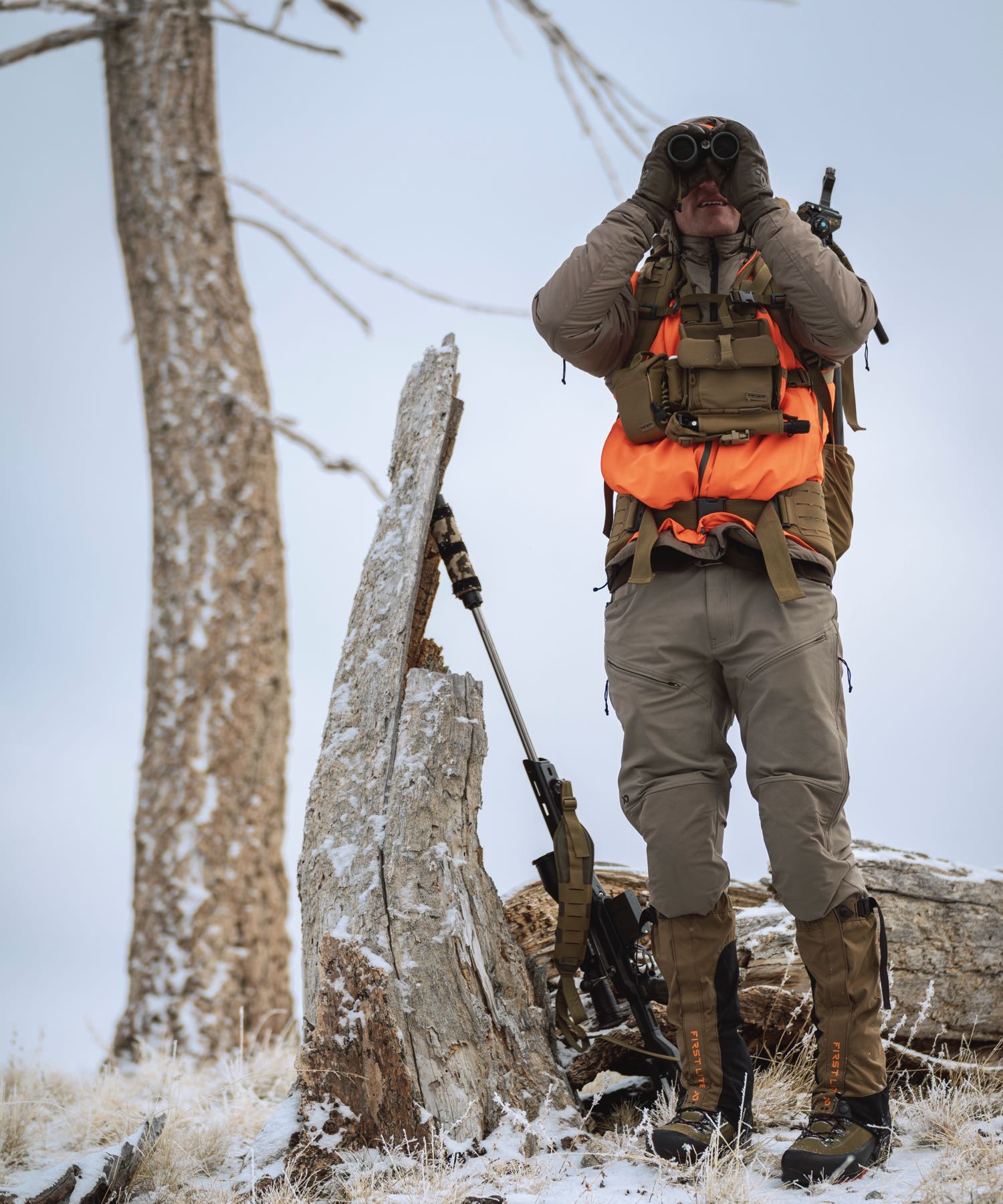Tan outdoor pants & brown First Lite gaiters on man scouting with binoculars in snow.