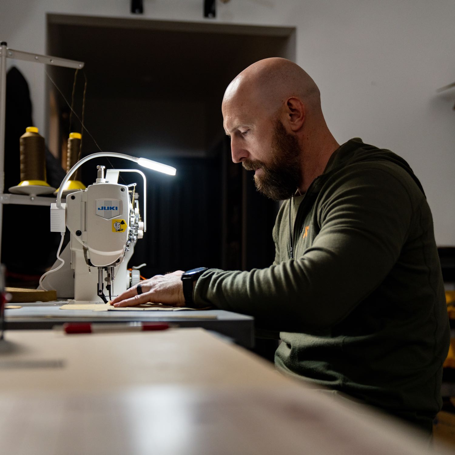 Man with beard in green shirt stitching tan material on JUKI sewing machine.