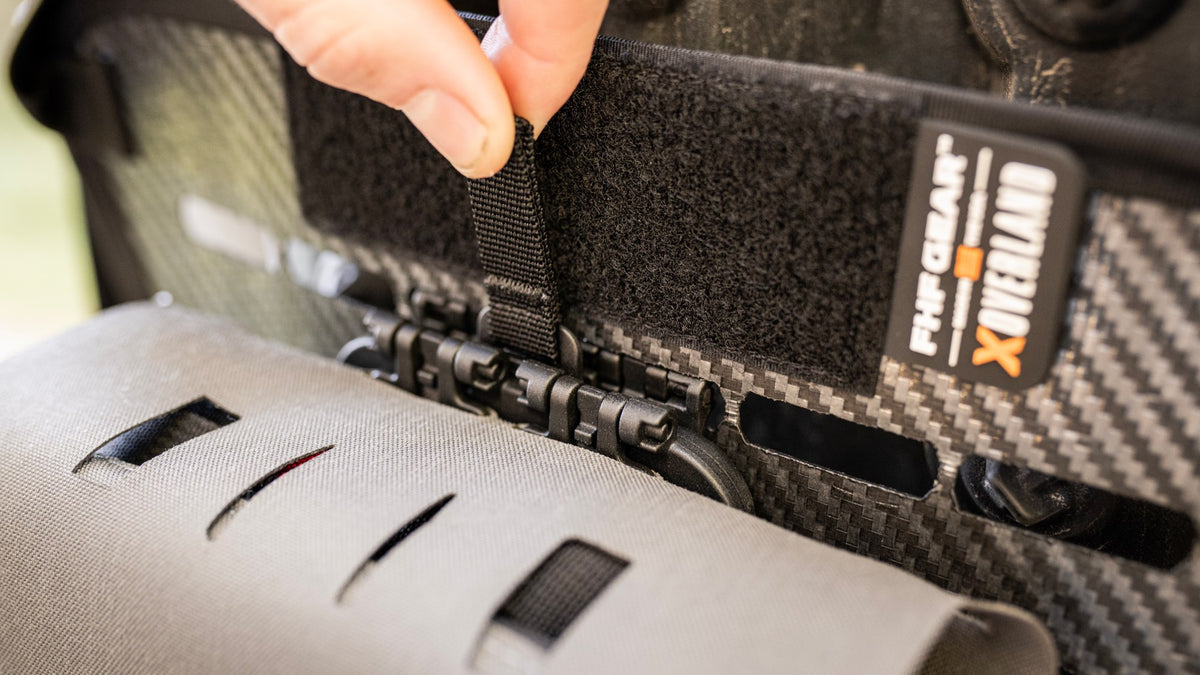 Close-up of a person's hand fastening a black hook-and-loop strap onto a black and gray carbon-fiber-patterned tactical vest or backpack, with a visible 'THE CLIMB X OVERLAND' brand patch.