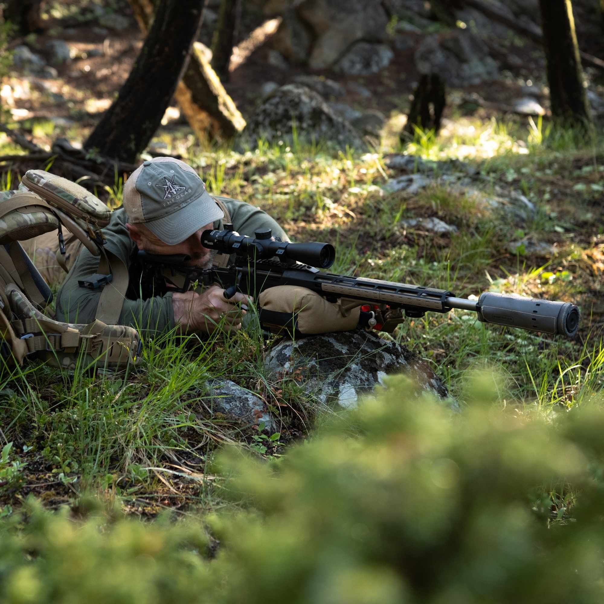 Hunter aiming rifle with black scope and  green suppressor cover on the ground in a forest